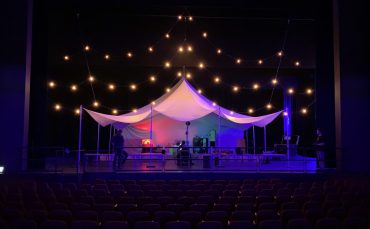 A tent, illuminated by strings of lights, sits on a theatre stage. Technicians, dressed in black, are at work.