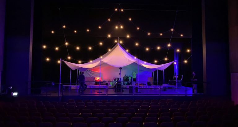 A tent, illuminated by strings of lights, sits on a theatre stage. Technicians, dressed in black, are at work.