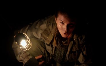 Person in a dark setting, partially illuminated by a hanging light, wearing a heavy jacket and looking upwards with a surprised expression.