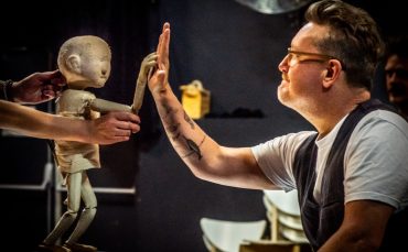 A man with glasses and a beard high-fives a puppet being manipulated by the hands of a person standing out-of-shot. The background includes chairs and a dark wall, creating a theatrical atmosphere.