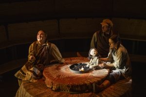 Three actors perform around a round table resembling a tree stump. One holds a puppet, and another looks upward, smiling. The scene is dimly lit, creating a warm, intimate atmosphere.